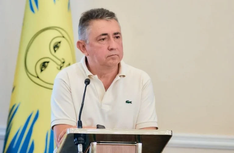 Чоловік у білому поло стоїть за трибуною з мікрофоном біля жовтого прапора