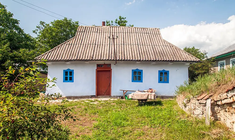 Біла сільська хата з синіми вікнами, черепичним дахом і дерев’яними дверима серед трави